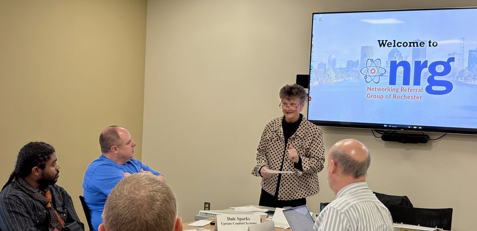 Lisa is speaking to NRG members with the NRG splash screen on the wall-mounted display behind her.