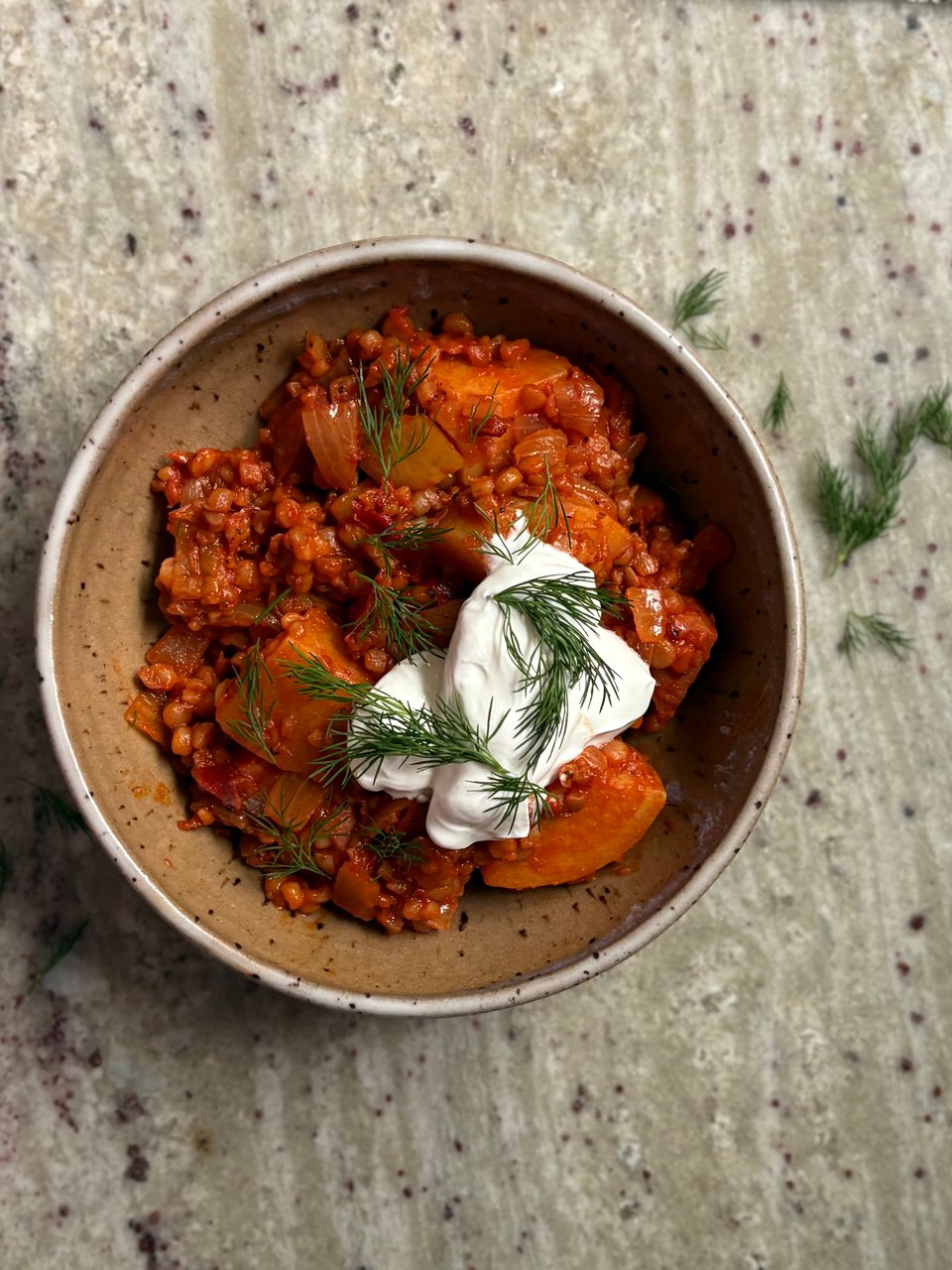 A ceramic bowl of red, sweet paprika-stained Hungarian-style shepherds stew topped with sour cream and dill.