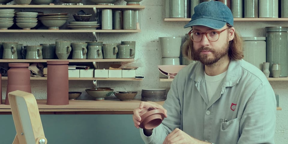 The image shows a potter in his studio holding a pot he has made.