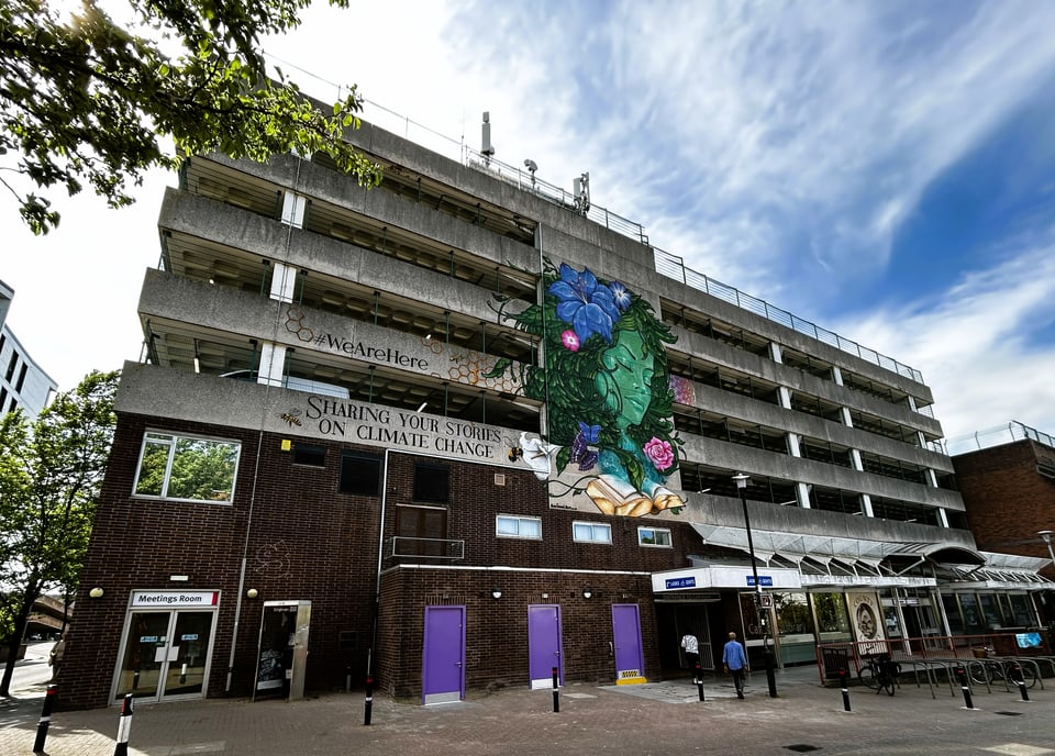 A large five storey concrete carpark with a library in the brick ground floor of it. The canopy of the library looks a lot like the whit 'wing' design common on Sainsburys branches in the 1980s. The solid concrete panels of the lift shaft for the car park have been painted with a mural of a woman made of plants emerging from the book she is reading. Text reads sharing your stories on climate change.