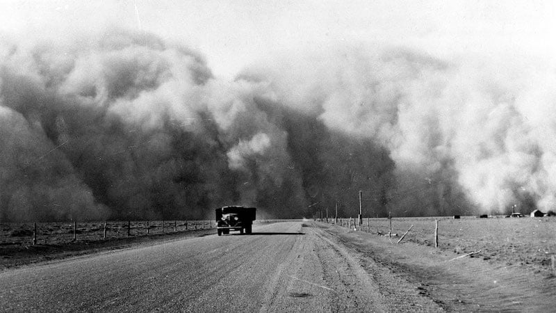 Black and white photograph showing an old truck driving away from a massive, billowing dust cloud. Road is straight and dirt. Landscape is flat.