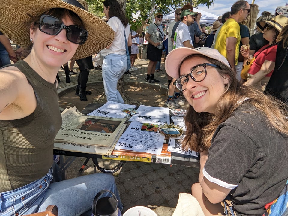 A picture of Lauren and Katie at a table filled with flyers, zines and stickers at the No Kings Demonstration