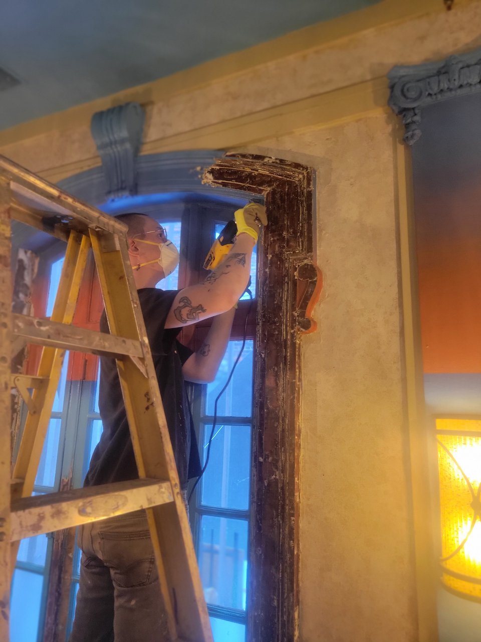 A friend scraping layers of paint off the windows with a heat gun. You can see the original island sunset colors on one half of the window, while the other half is dark exposed wood.