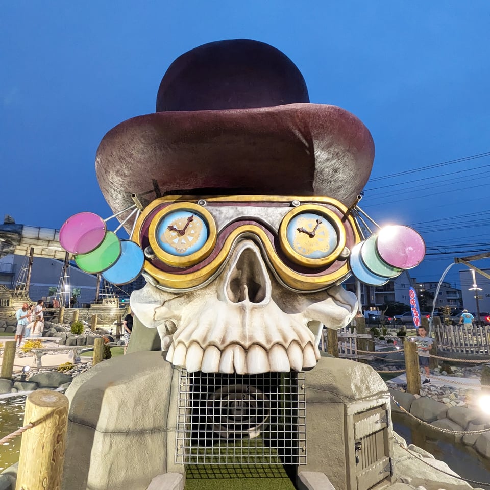 A giant fiberglass skull wearing a top hat and weird glasses with clocks on each lens. Its teeth rest on the grate covering the final hole at the mini golf course.