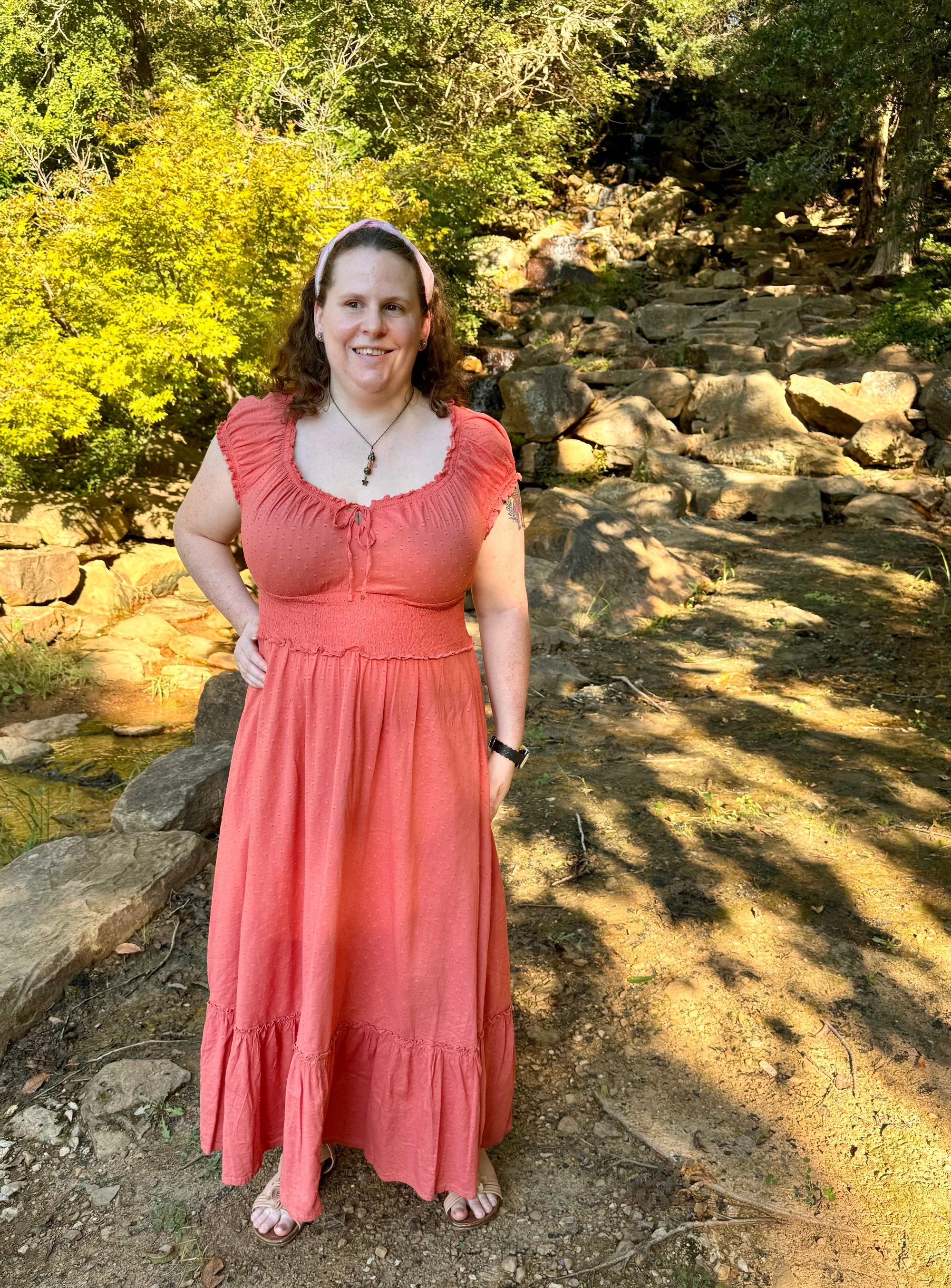Daisy, a fair-skinned woman with freckles, standing in a wooded area with her hand on her hip and a small waterfall off to the side, wearing an ankle-length coral-colored dress with cap sleeves, a scoop neckline with a tie, a cinched waist, a flowing skirt with a ruffled hem, and little textured dots all over, a shiny pink fabric headband holding back her auburn curls, a necklace with a vial of flower petals and a dangling flower charm, pink flower earrings with stones in the center, and tan strappy sandals with 3D flowers on the top that may or may not be visible