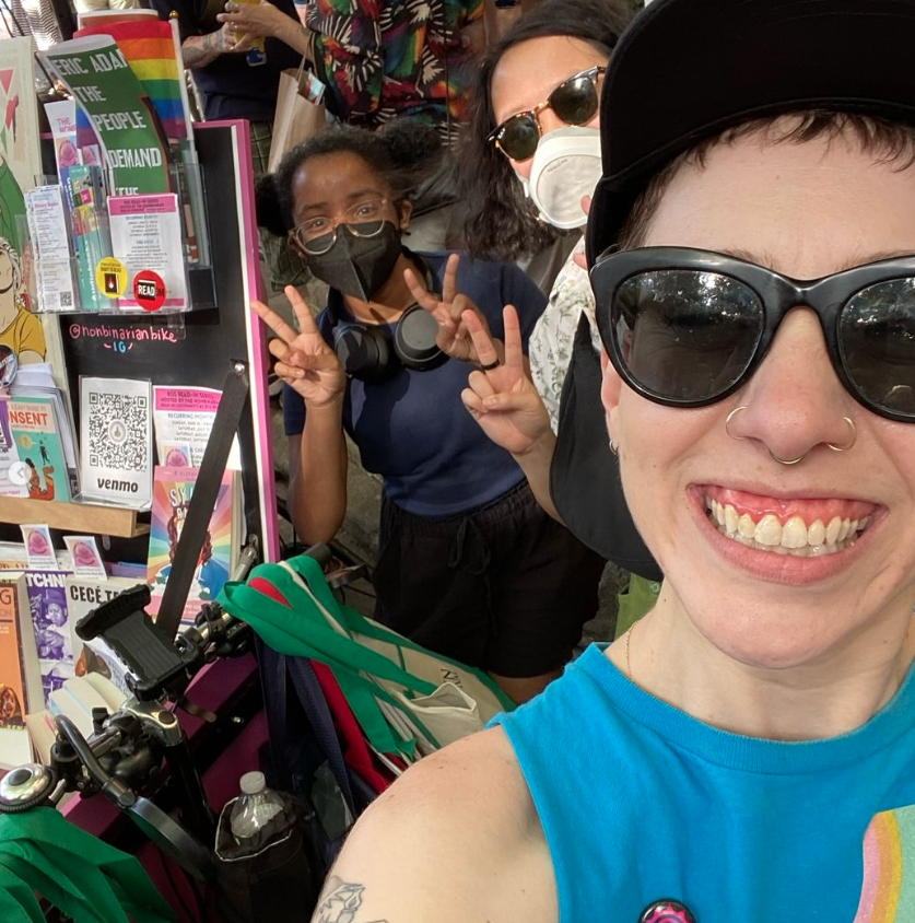 The Nonbinarian Book Bike founder K. Kerimian poses with volunteer coordinator Alyssa Lo, volunteer Marlowe, and the Book Bike at Brooklyn Pride 2024