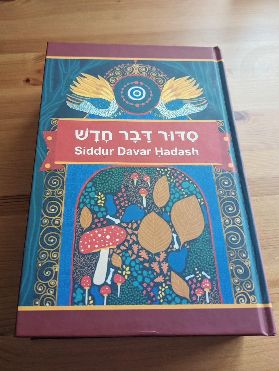 A physical copy of Siddur Davar Ḥadash resting on a table. The cover features forest motifs of leaves, mushrooms, soil, and bones.
