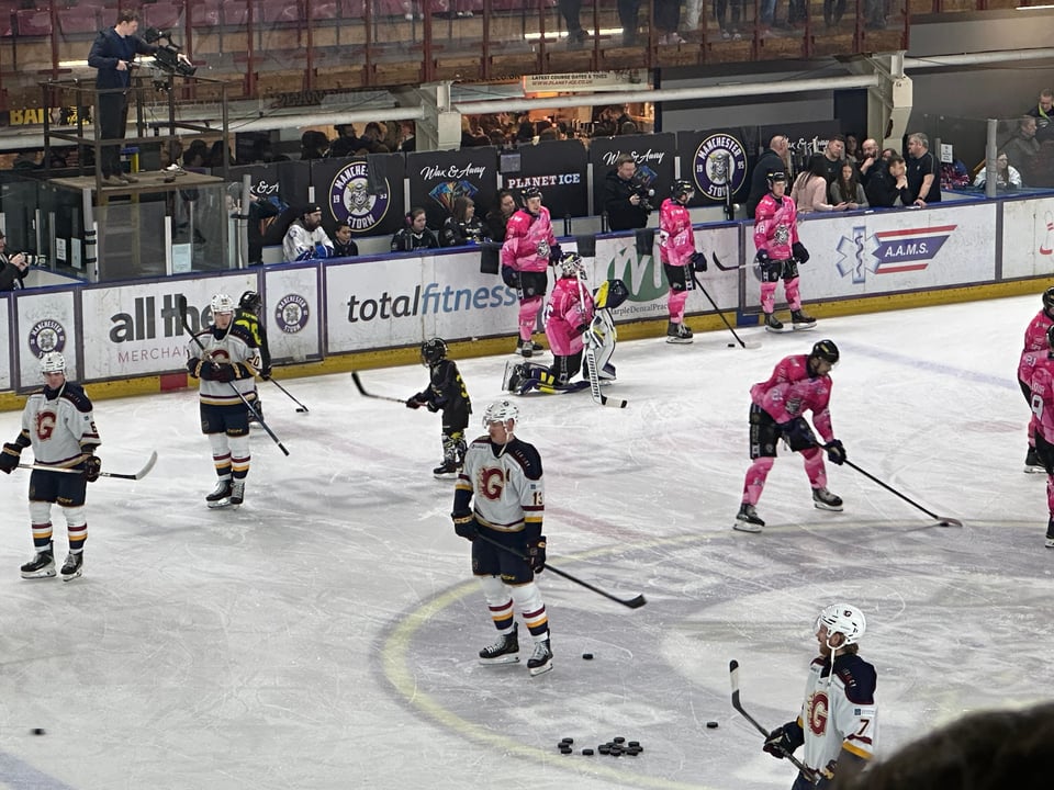 A number of ice hockey players on a rink, some in pink and black, others in white and black. Three children dressed in black.