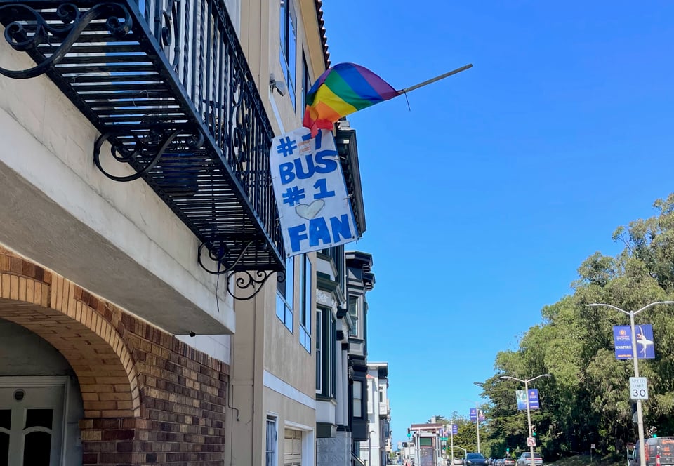 a sign that reads "#7 bus, #1 fan" hangs out of a window near the #7 bus stop, under a rainbow pride flag