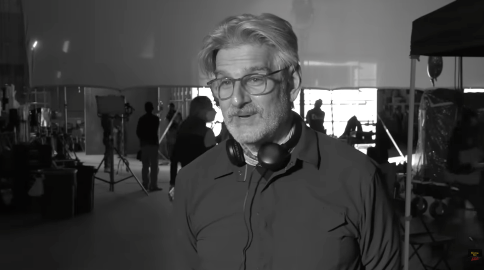 Black and white photograph of a grey-haired man being interviewed on camera. He has a pair of headphones round his neck. In the background we see a film crew setting things up.
