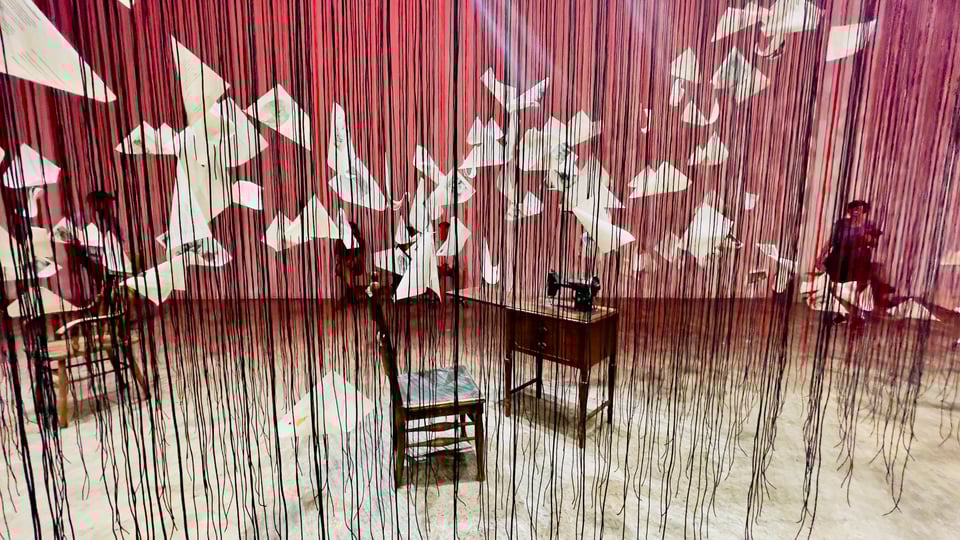 Thin, red and black strings hanging from the celling set the scene for sheets of paper seemingly floating in mid-air, an old wooden chair and and old-timey manual sewing machine on its wooden stand