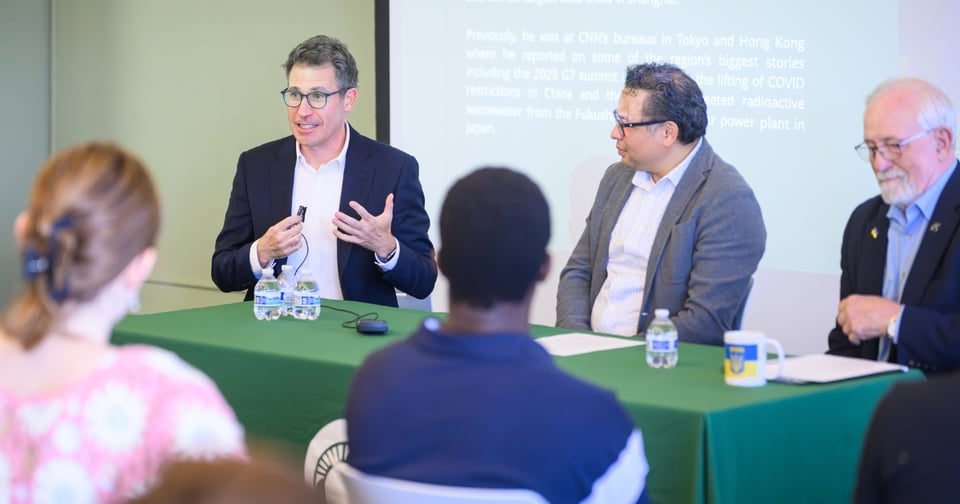Three men seated together at a table with a green tablecloth. The man on the left is speaking into a small microphone and gesturing with his hand. The two men on the right listen attentively. Audience members are visible in the foreground, suggesting a public lecture setting.