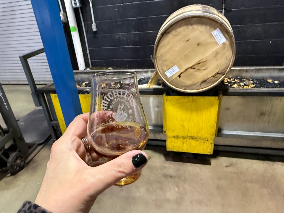 glass of Michter's rye held aloft near barrel being dumped into filter trough