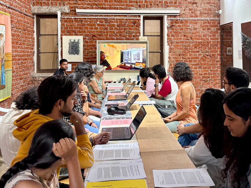 People seated at a long linear table with four laptop computers on one side and a table runner print in brown on the other side. The table also has sheets of paper in pink and white, and is helmed by Maya Sharma at the end and a television unit behind her, playing a video with a view of a similar tabletop setting from the Just Futures Co-lab. The exposed brickwork of the walls is contrasted with visual art work on each of the longer sides of the gallery.