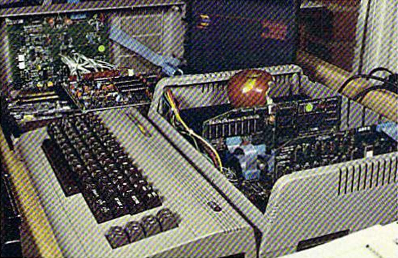 A Commodore 64 plugged into a Spartan machine, sitting on a desk. The Spartan has its top off and an apple sitting on top of its exposed power supply.