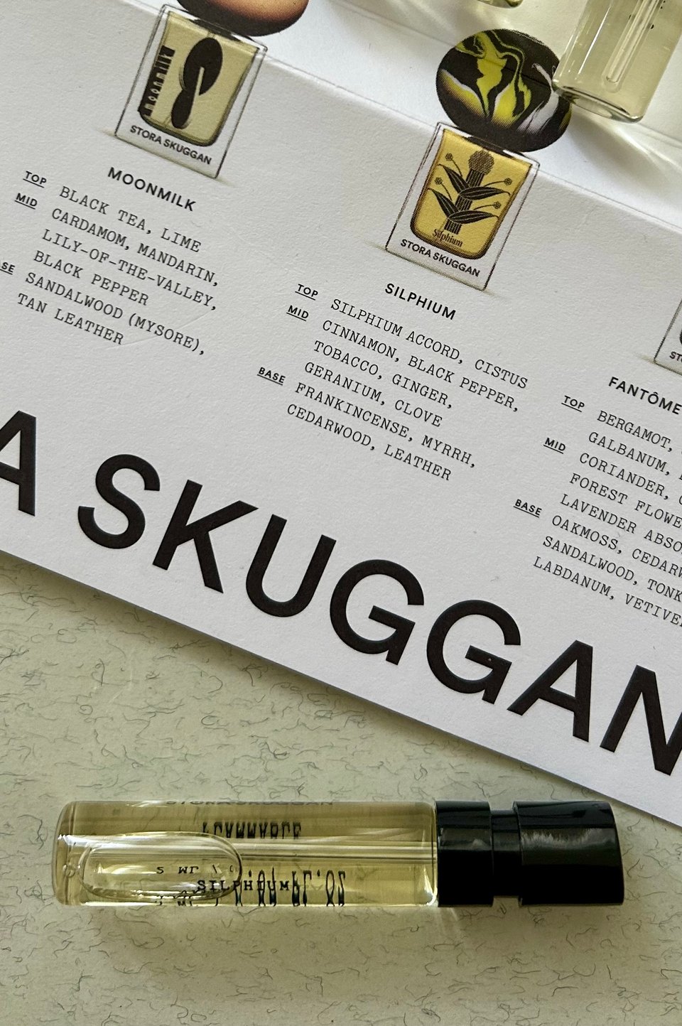 A glass vial of Silphium by Stora Skuggan perfume is resting on a cream with grey flecks notebook. There is Stora Skuggan branded packaging behind, listing the notes you may be able to detect in the perfume. Image by Rowan Ambrose.