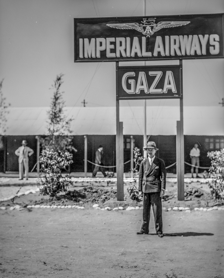 A man in a double-breasted suit and a hat standing in front of a sign that says Imperial Airways Gaza