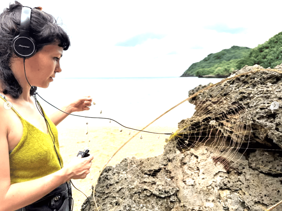 A profile of Audrey Briot wearing headphones and standing on a beach holding her work for her project Ocean of Waves.