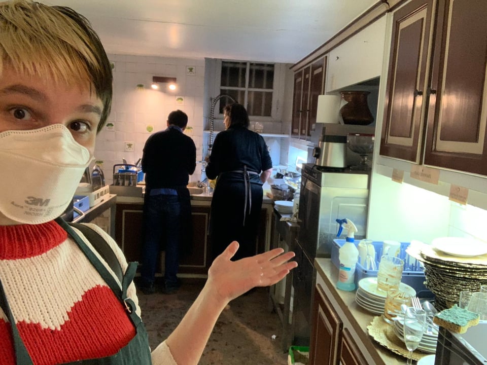 A person with a mask on shows the amount of dishes needing to be cleaned while two people in the back of the kitchen are busy washing the dishes.