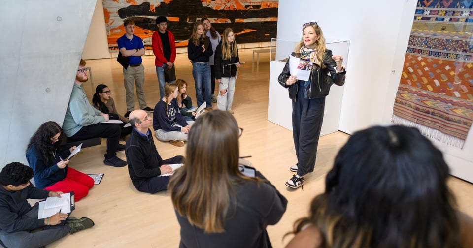 A JMC professor stands in a modern art gallery, holding papers and gesturing as she speaks to a group of students seated on the floor. The attendees, including teenagers and adults, are attentively listening, some taking notes, while colorful artworks adorn the walls around them.