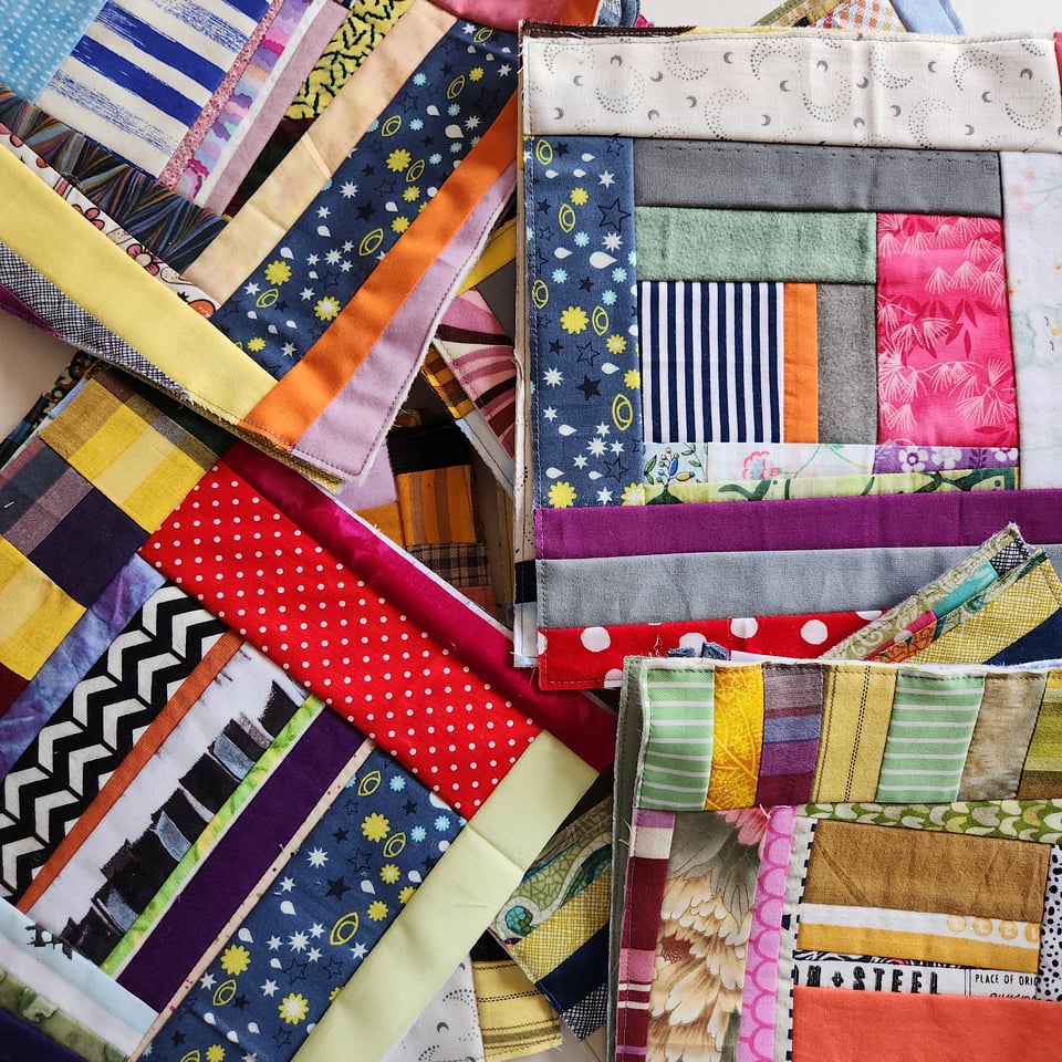 Top-down photo of many patchwork potholders in a pile. They include fabric in many different colors and patterns.