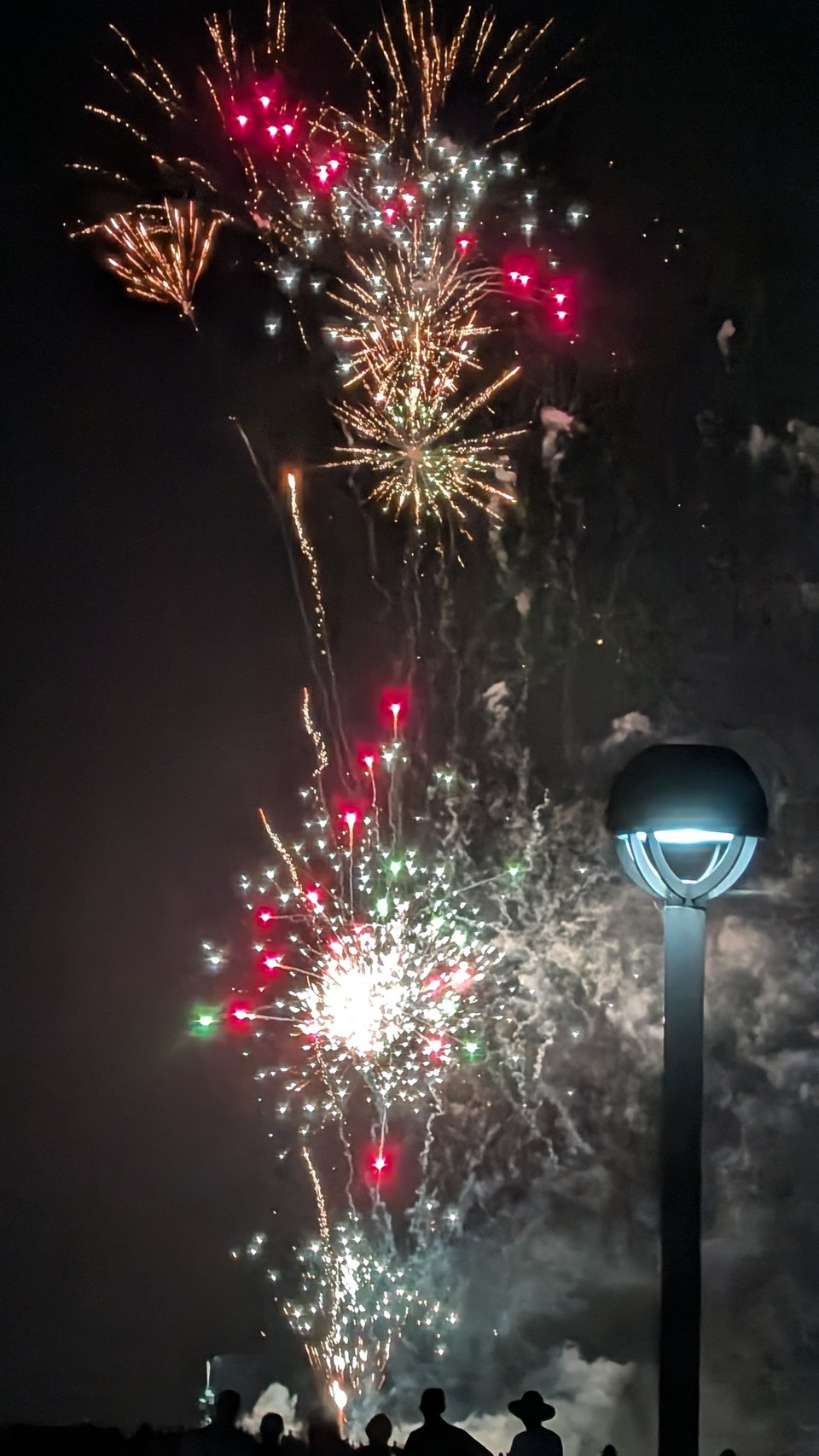 Fireworks explode against the night sky. Red, white, orange pops of color. Silhouettes of spectators can be seen at the bottom of the photo. Lots of smoke too.
