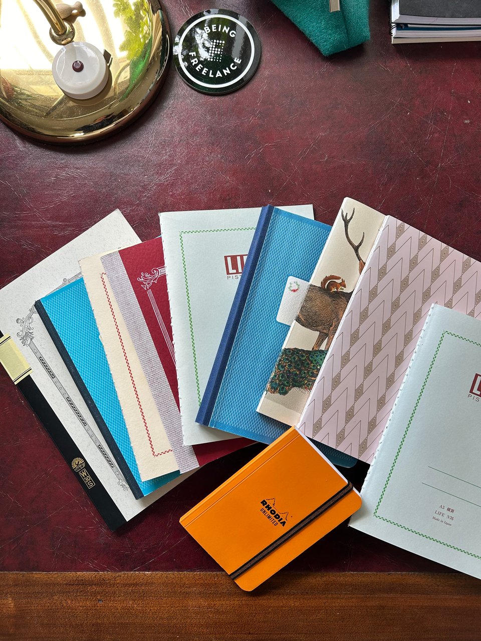An array of patterned notebooks are fanned across a dark red leather topped desk. Image by Rowan Ambrose.