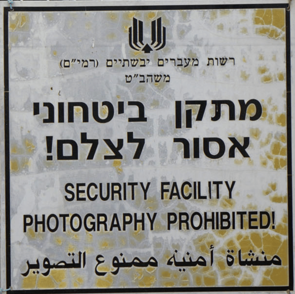 An image of a sign with writing in Hebrew and in Arabic, and English words that say, in all caps, SECURITY FACILITY - PHOTOGRAPHY PROHIBITED!