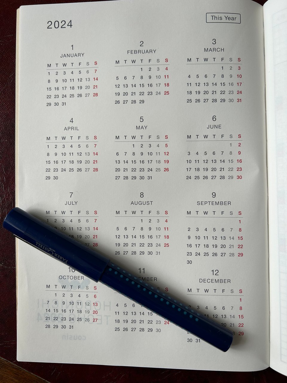 A page of a desk planner showing a 2024 calendar by month. There is a dark blue fountain pen lying at an angle on the page.