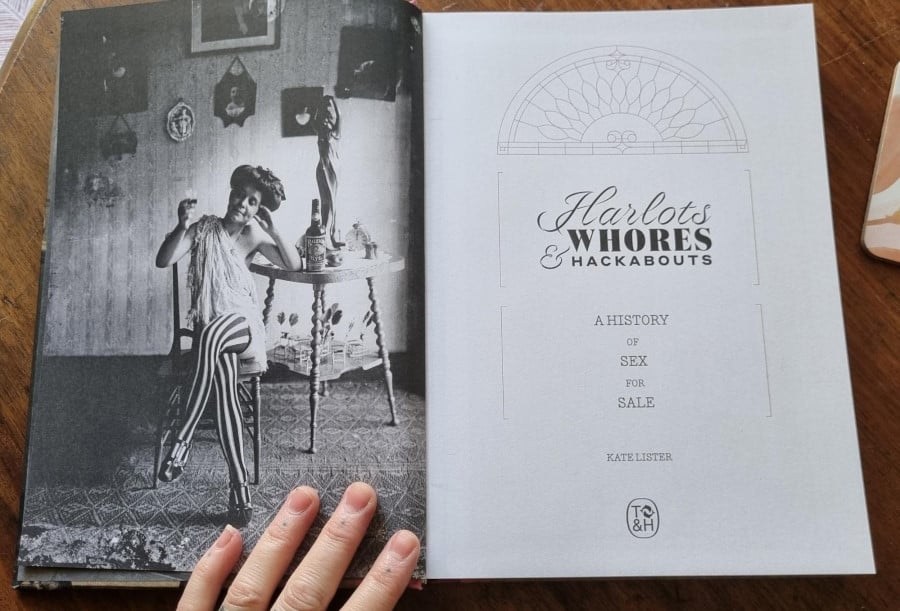Photograph of the title page of Harlots, Whore & Hackabouts, the preceding page on the left shows a black and white photo of a woman wearing long black and white striped leggings, leaning on a bar table, looking longingly at the drink in her hand.