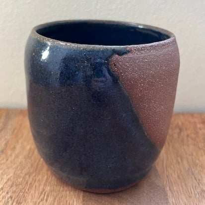 A simple cup made of red clay. It's been glazed in dark blue, but a diagonal section has been left bare with the clay showing.