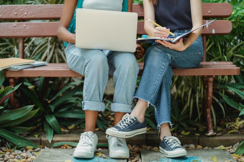 Image of two people sitting on a bench while writing in an open notebook and a laptop.