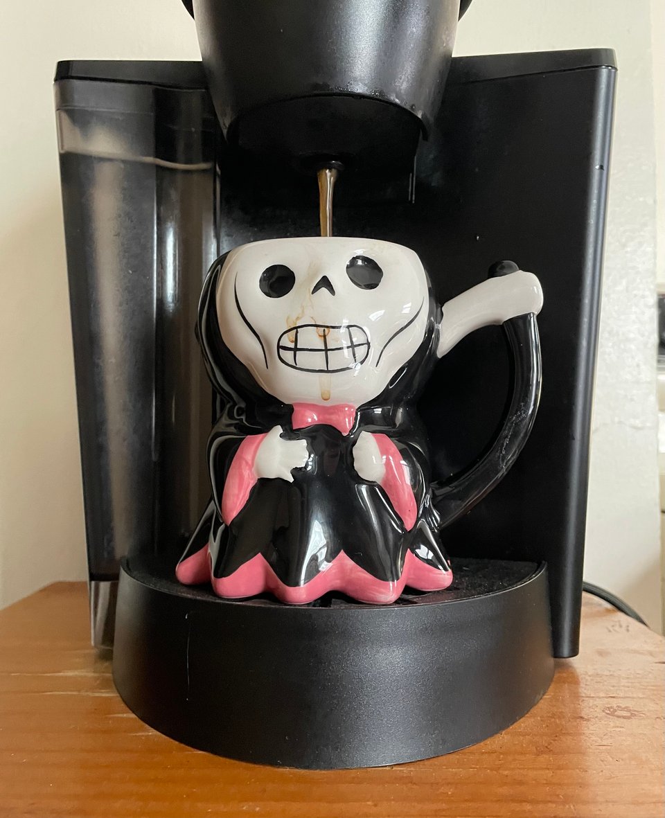 A coffee mug shaped like a cute version of the Grim Reaper grimaces as coffee from a Keurig machine drips into its head