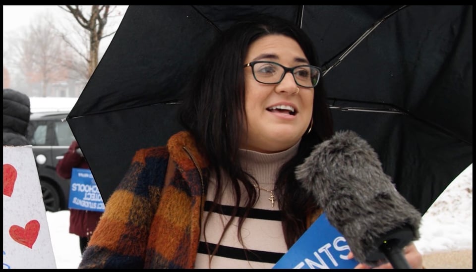 A protesting woman in light winter gear and umbrella speaks into a microphone with a massive fuzzy wind baffle.