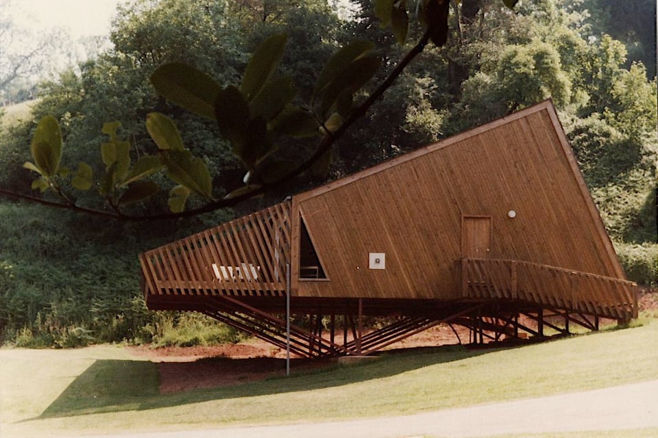 A wooden holiday lodge in a long ago summer sun. It's a wedge resting on a metal framework over sloping ground. At the front, there's a balcony. The rest of the lodge is a solid wall of diagonal wooden cladding with nothing but a triangular window and a regular door set into it. A walkway leads to the door.