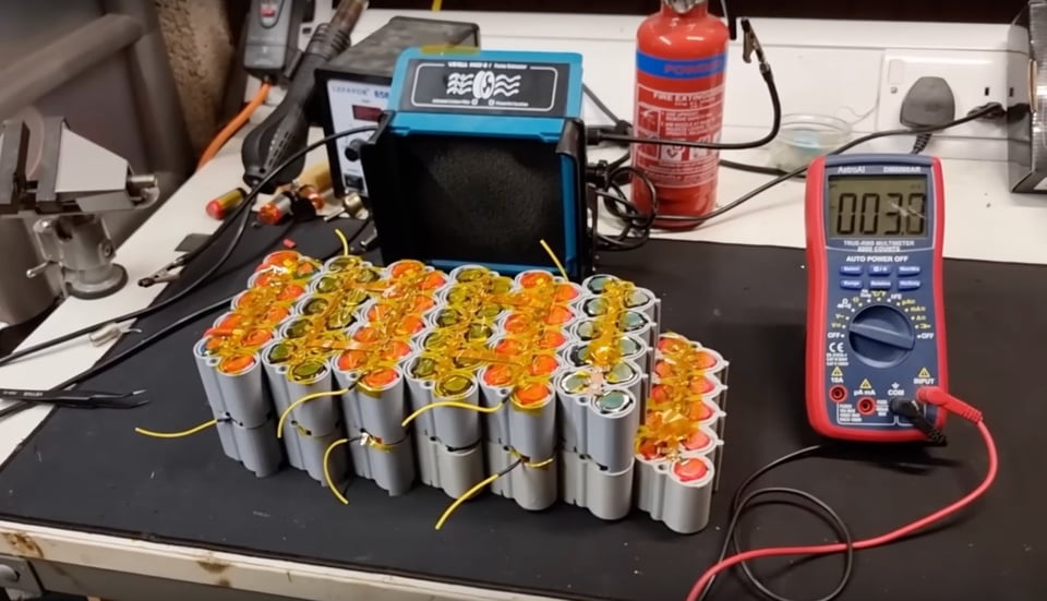 A photo of a workbench on which sits perhaps 120 small batteries — in size and shape very much like AA batteries — all collected together and standing upright inside a DIY plastic gray casing. There are several wires sticking out from the apparatus. Directly to the right of the apparatus is a multimeter, and in the background there appears to be a soldering iron, and a red canister of unknown function.