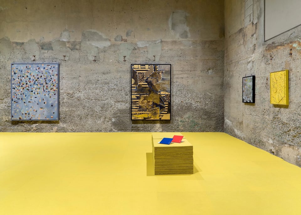 View of abstract paintings hanging on two walls of a gallery with concrete walls and bright yellow carpeting