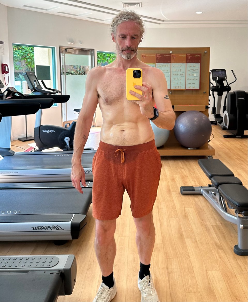 Full-body mirror-selfie of a white man in rust-colored sweat-shorts & no shirt in a hotel fitness center. He just ran 30 minutes and looks pretty good