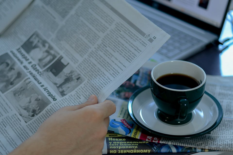 A hand holding a newspaper, next to a cup of coffee.  Photo by Anna Keibalo on Unsplash