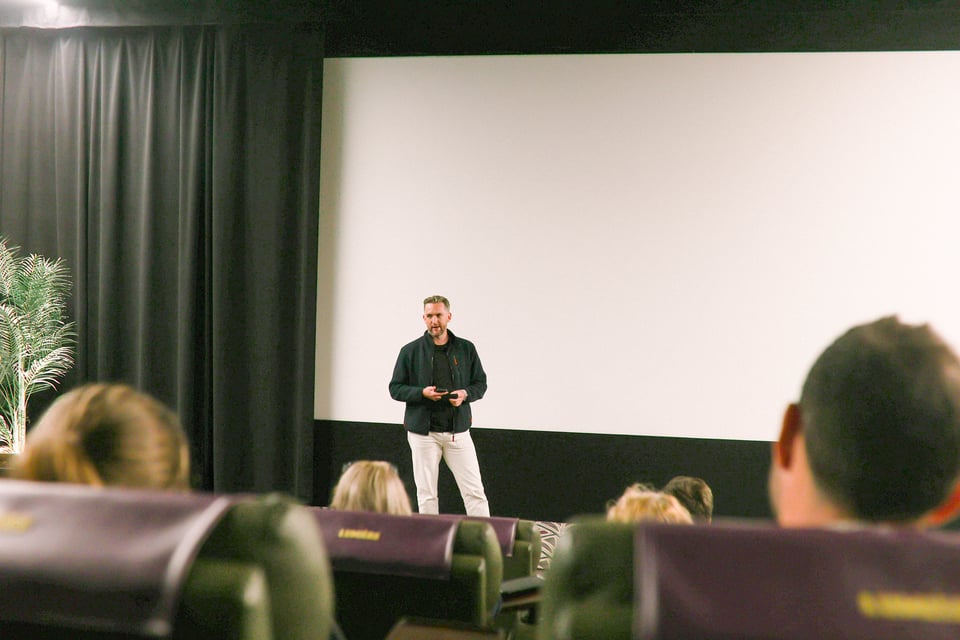 Filmmaker, Harry Knight, is stood in front of an audience in a cinema. He's wearing white trousers, an open navy jumper and a black t-shirt. He's standing in front of a blank white screen with a drape in the right third of the frame. In the foreground we see backs of audience members heads.