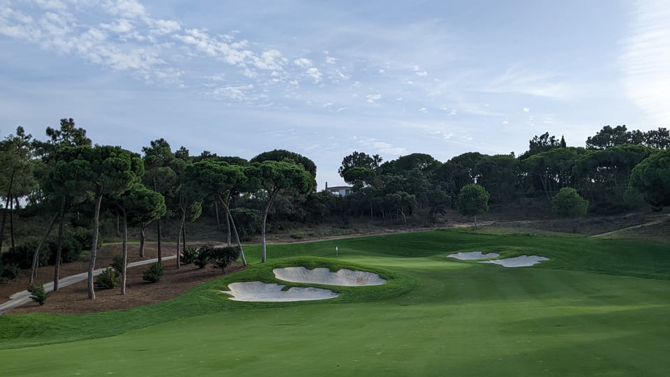 18th hole green on Quinta do Lago North, Portugal, from the downhill fairway. Large greenside bunkers filled with white sand, framed by umbrella tree-lined path.