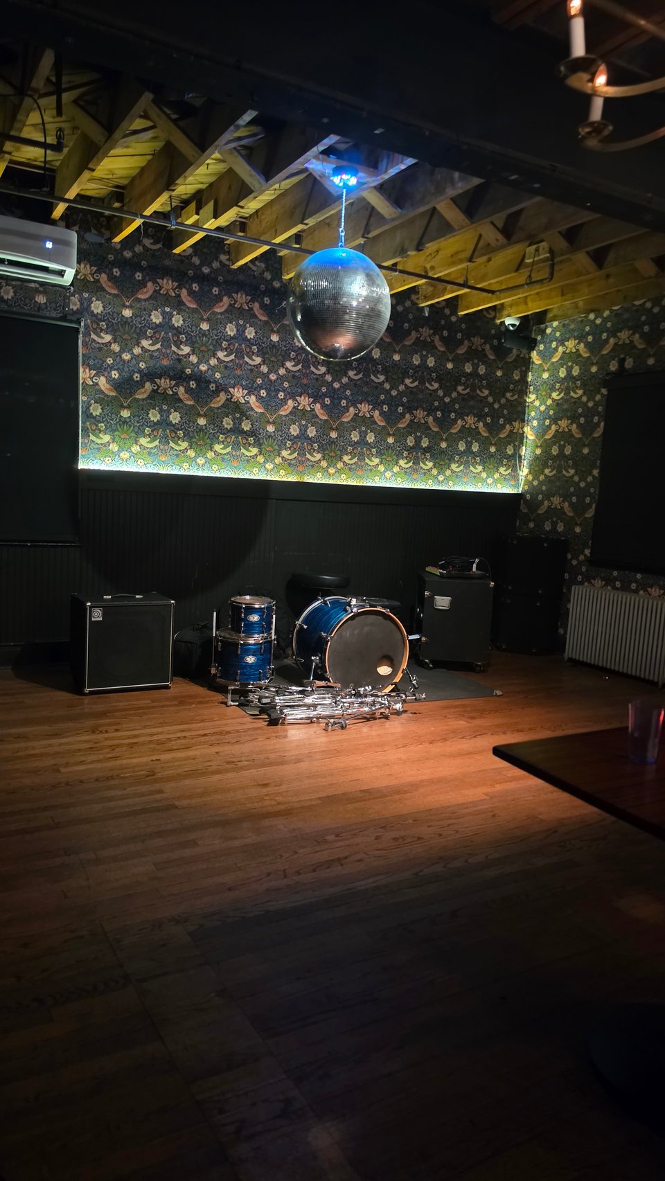 A dimly lit music venue with a blue sparkle drum kit and amplifiers set up against a wall with floral bird-patterned wallpaper, a mirrored disco ball hanging from exposed wooden ceiling beams, and a wooden dance floor in the foreground.