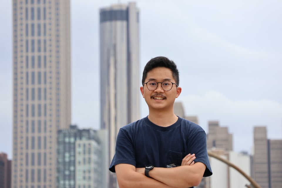 Alex, an Asian man with short black hair, glasses, a navy shirt, and a smart watch, poses in front of the Downtown Atlanta skyline