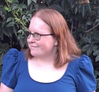 Headshot photograph of a white woman with pale, freckled skin, red hair, and glasses. She is wearing a blue dress and smiling while she looks off to her right.
