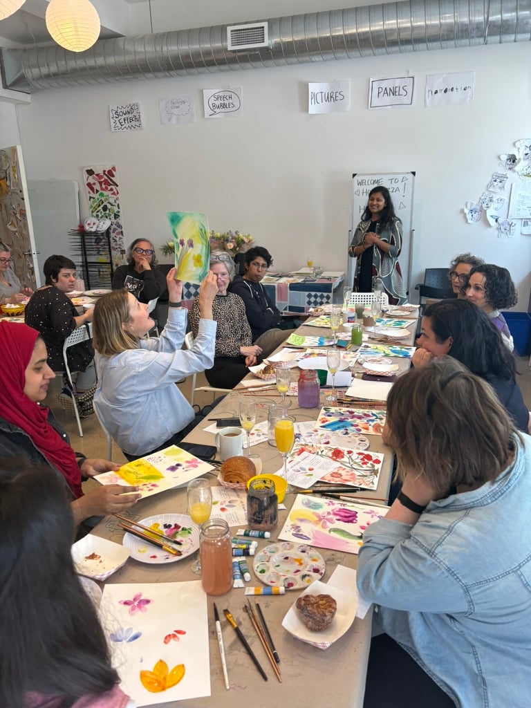 Women around a table making watercolour paintings on paper; one is holding hers up for the others to see.