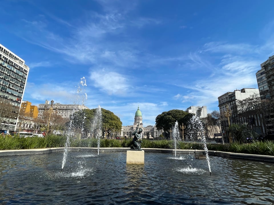Fountain in the plaza de los dos congresos with a small statue in the middle of four water spouts shooting into the air. In the far background the congress building and its dome are visible.