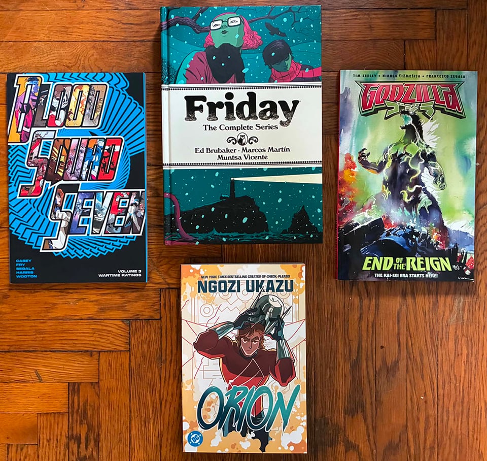 A photograph of the books Blood Squad 7 volume 3, Friday, Godzilla: End of Reign, and Orion on my floor.
