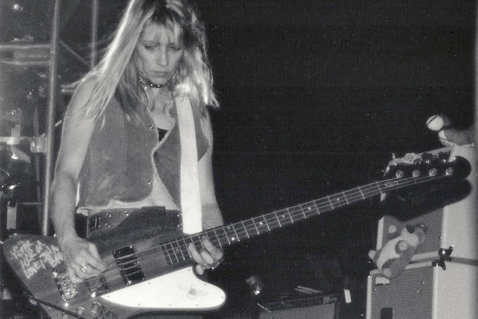 A black-and-white photo of Kim Gordon, the bassist for sonic youth, from the waist down. She is playing a bass that has a roughly thunderbolt shape; it's made of brown natural wood with a white pickguard, and has some white handwriting on the far end. She is dressed in a leather vest and is looking down at the fret board.