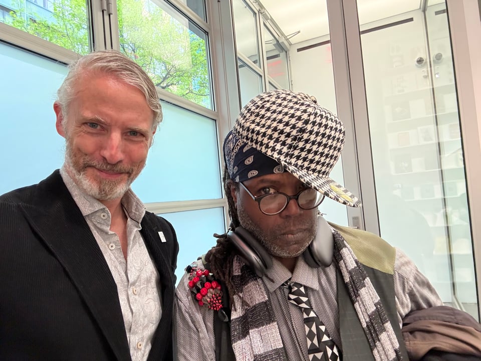 Double-selfie photo of white man in black corduroy suit and paisley dress shirt shirt standing beside a black man wearing a whole ton of black-white-gray clothes