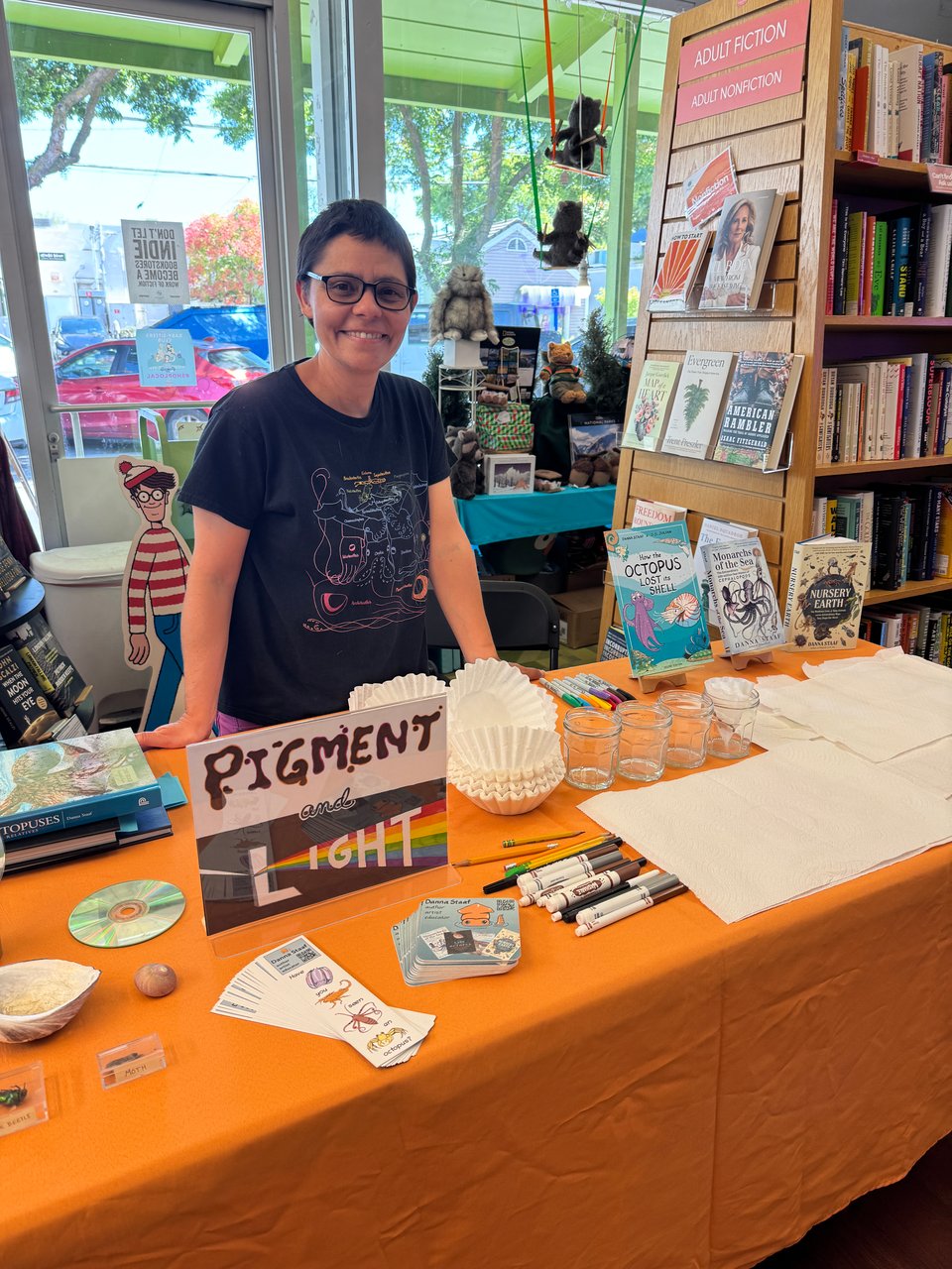 Photo of me (Danna Staaf) standing smiling behind a table in Linden Tree bookstore. The table has an orange tablecloth, a Pigment and Light sign that I drew, several of my books, and all the materials for marker chromatophy laid out. I am wearing a my rainbow shirt of comparative squid sizes. Photo taken by the marvelous Rie Neal.
